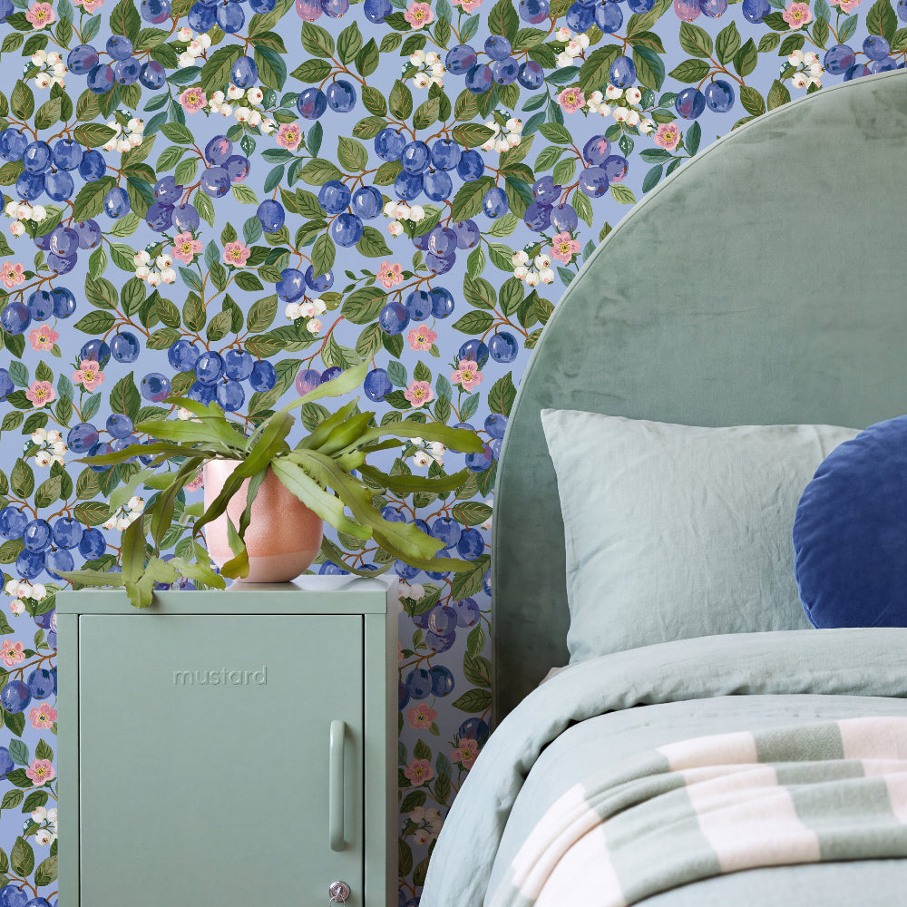 A styled bedroom corner featuring a light blue floral wallpaper with blueberries, green leaves, and small pink and white flowers. A mint green nightstand with a potted cactus sits beside a bed with a matching mint green headboard, white and blue pillows, and a striped blanket.