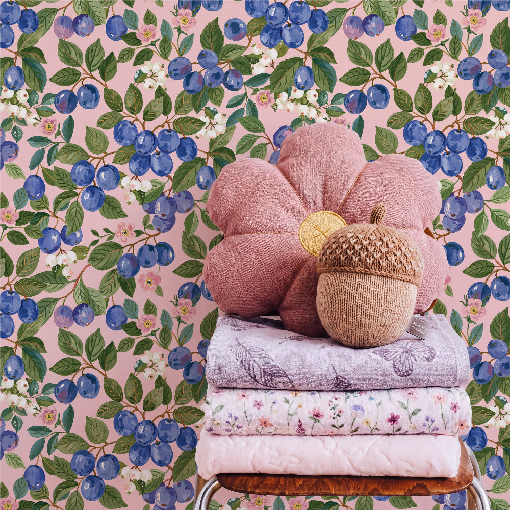 A styled product display featuring a dusty pink flower-shaped pillow with a gold coin accent, stacked on folded patterned quilts next to a woven acorn pillow, set against a pink watercolor blueberry wallpaper background.