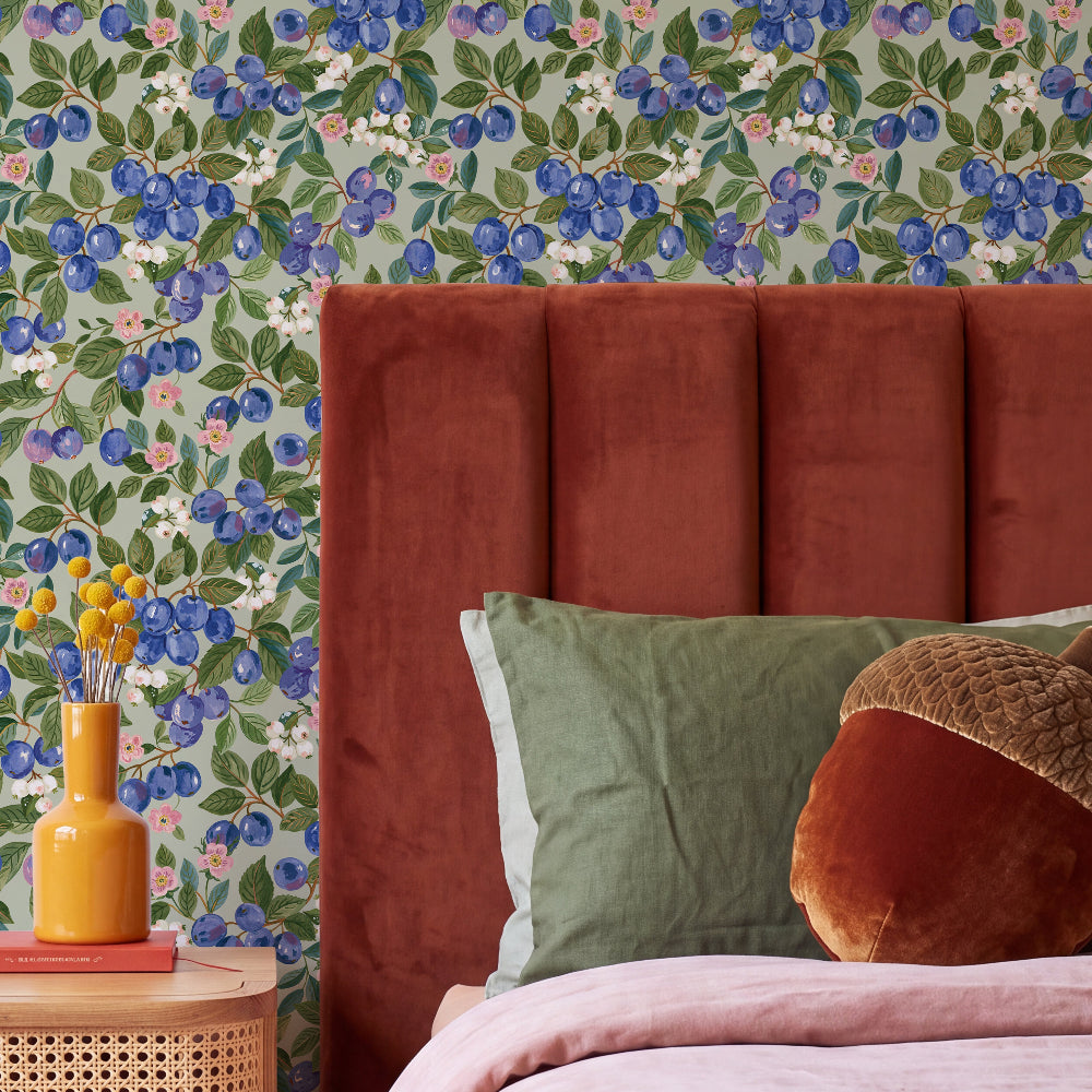A styled bedroom scene featuring a tall, ribbed headboard in rust red velvet, layered with sage green and brown pillows, set against a light green wallpaper with a blue berry pattern.