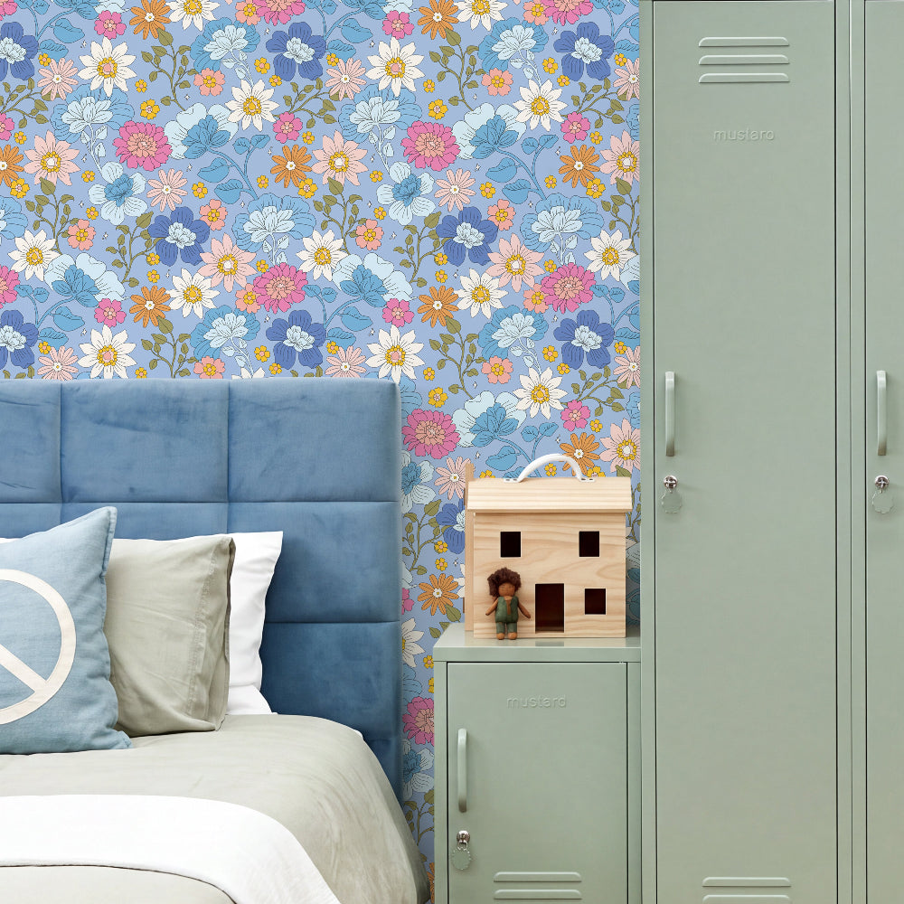 A styled bedroom corner featuring a blue tufted bed headboard and bedding, a floral wallpaper panel in blue, pink, white, and yellow, a sage green metal locker, and a small wooden house toy on a bedside table.