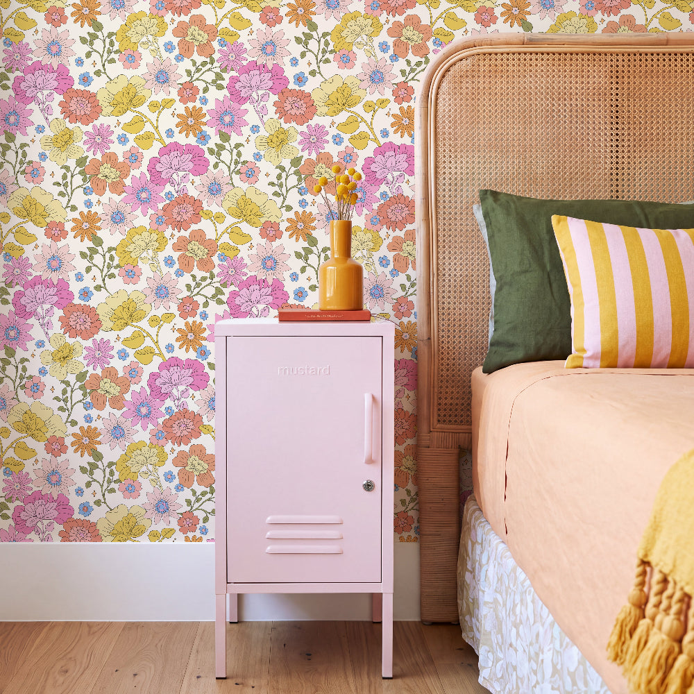 A styled bedroom corner featuring a light pink metal nightstand with a floral wallpaper background, a rattan headboard, and colorful pillows.
