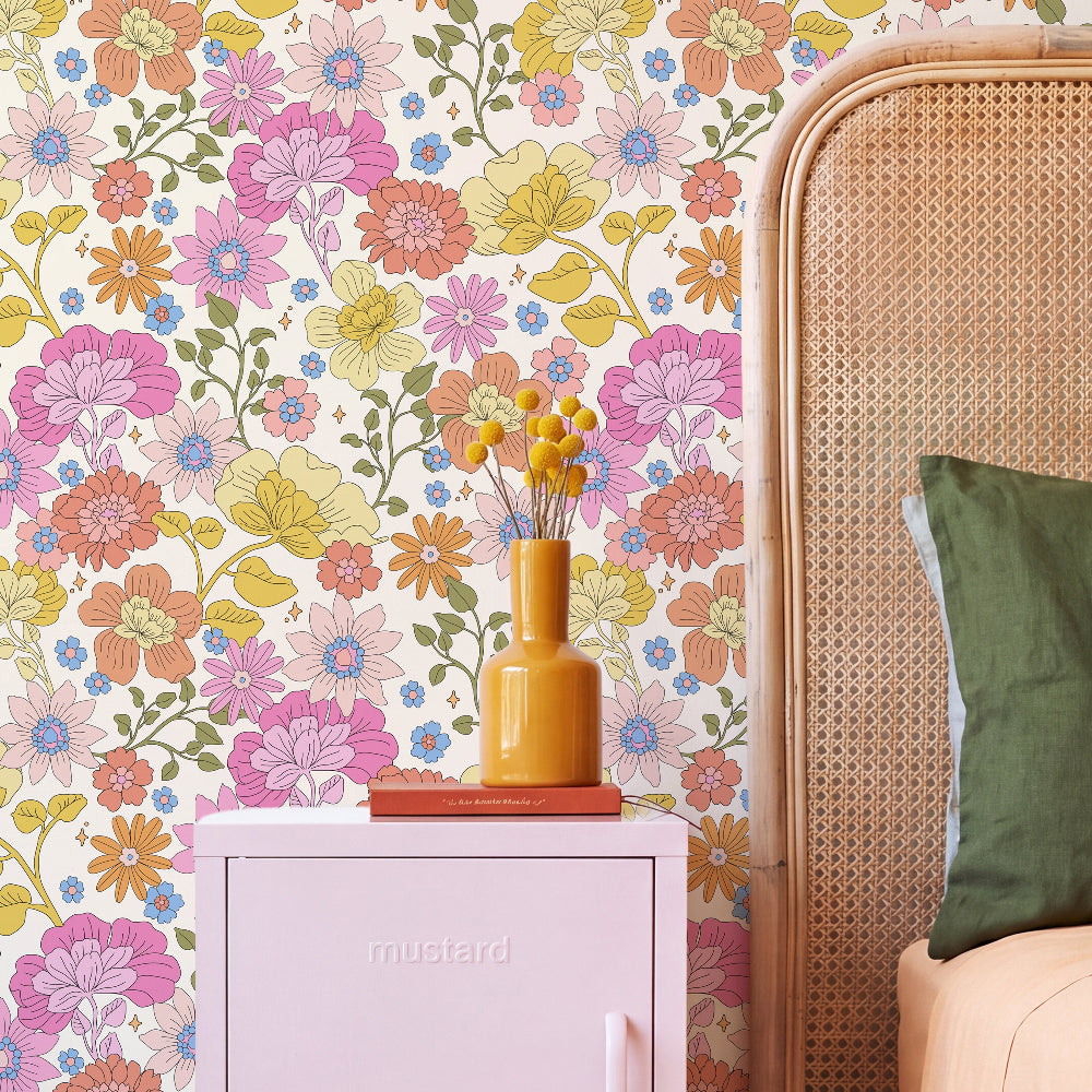 A styled bedroom corner featuring a wall covered in colorful retro floral wallpaper. A light pink side table with a yellow vase sits in front of the wall, next to a woven rattan headboard and a green pillow.