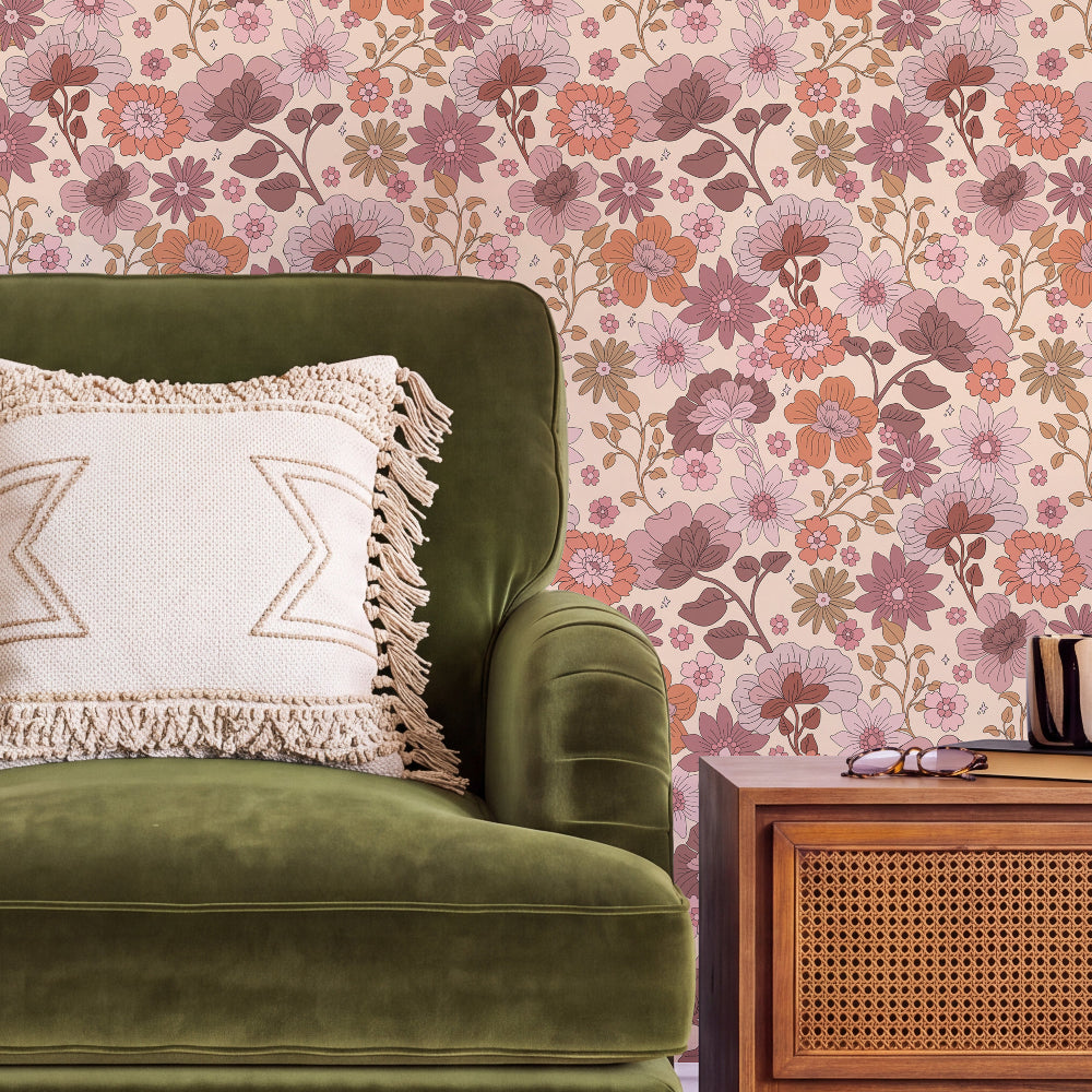 A styled interior scene showing a green velvet armchair with a cream fringed pillow, next to a wooden side table, in front of a pink and mauve floral wallpaper background.