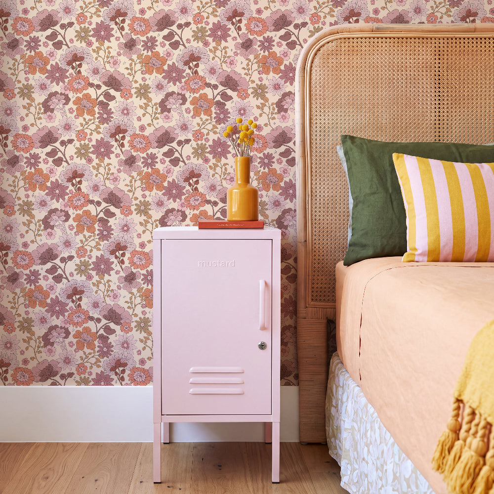 A styled bedroom corner featuring a light pink metal cabinet with a floral wallpaper background, a wooden cane bed headboard, and colorful pillows.