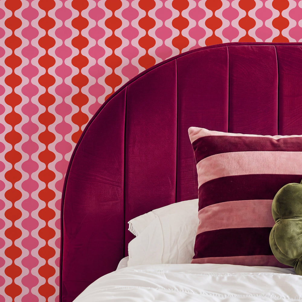 A close-up of a bed featuring a large, curved magenta velvet headboard, white bedding, a striped pink and burgundy pillow, and a green plush toy against a bold pink and orange geometric patterned wallpaper.