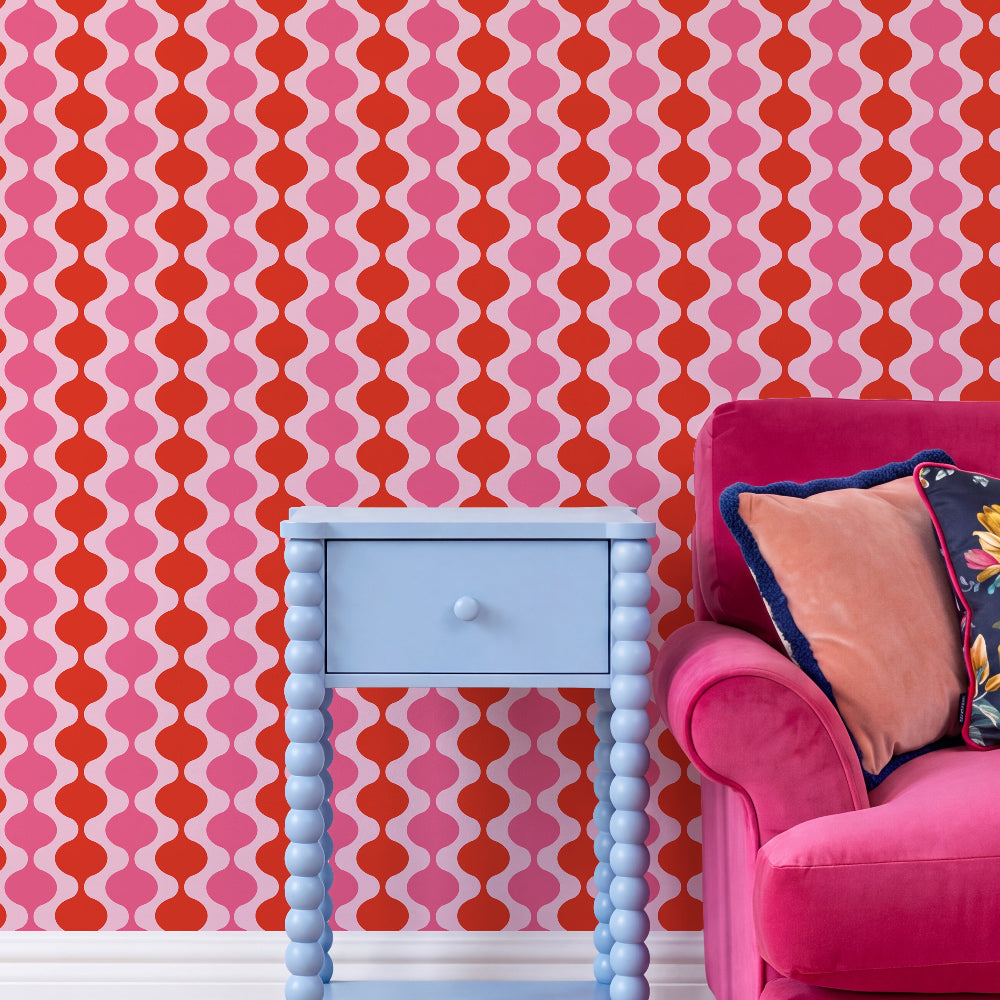 A styled interior corner featuring a bold pink, red, and orange geometric patterned wallpaper, a light blue single-drawer bedside table, and a bright pink velvet armchair with decorative pillows.