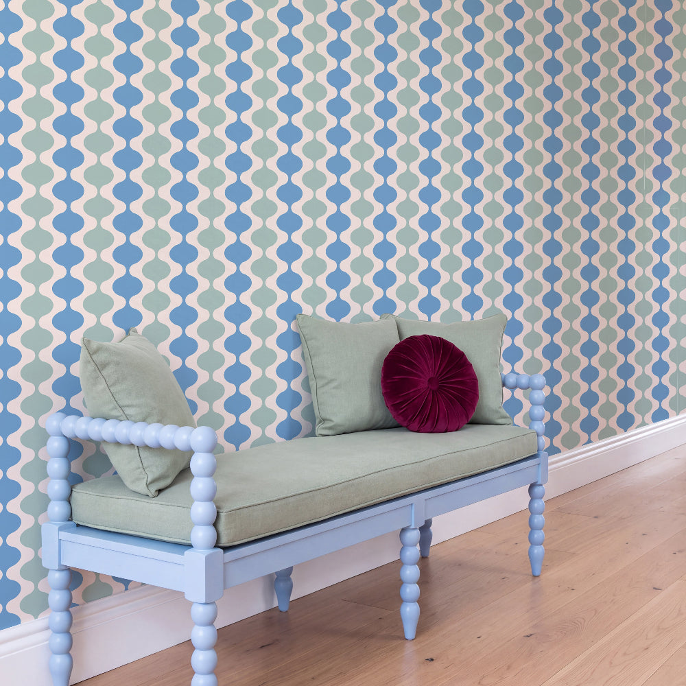 A light blue wooden bench with a sage green cushion and pillows, set against a blue and green geometric patterned wallpaper and light wood flooring.