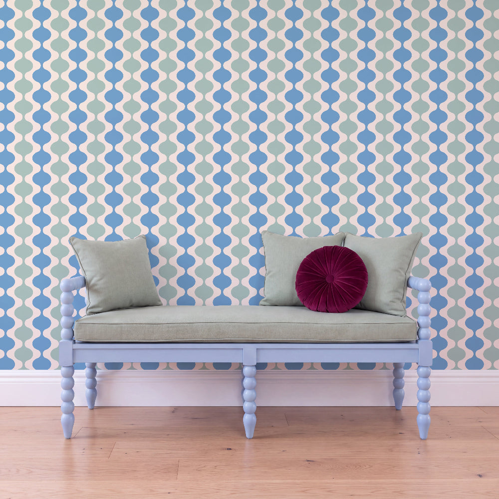 A light blue wooden bench with a pale green cushion sits against a wall covered in a repeating blue and cream geometric pattern. Two matching pale green pillows and a round deep magenta accent pillow rest on the bench, with a light wood floor below.