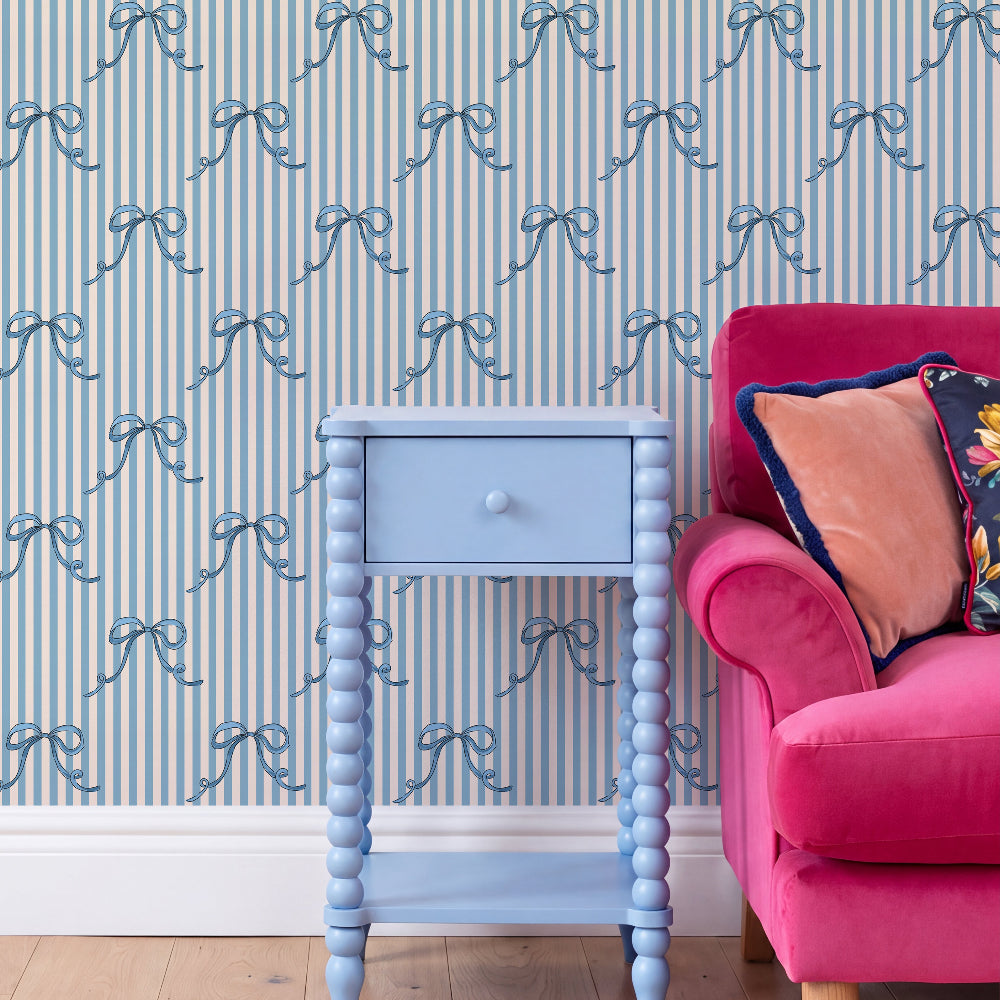 A styled interior scene showing a light blue bead-frame nightstand in front of blue and white striped wallpaper with blue ribbon motifs, next to a bright pink armchair with decorative pillows.