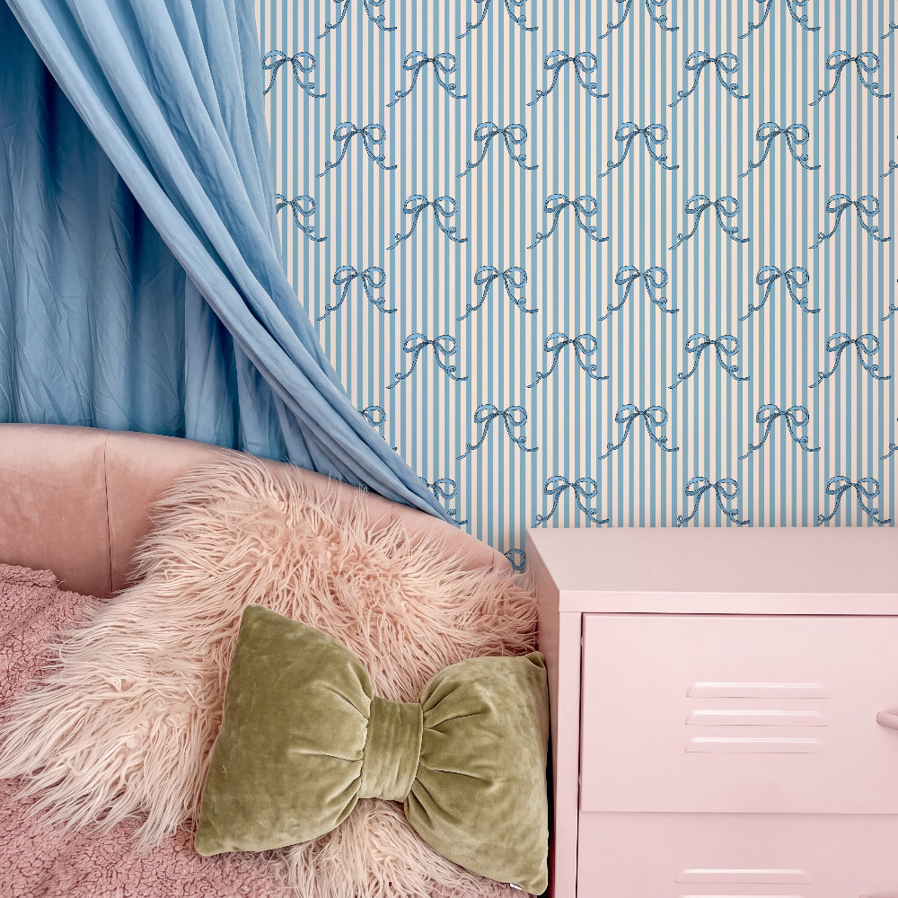 A styled bedroom corner featuring a light blue and white striped wallpaper with a repeating bow pattern, a dusty blue curtain, a blush pink bed frame, a fluffy cream faux-fur pillow, and a matching green velvet bow accent pillow.