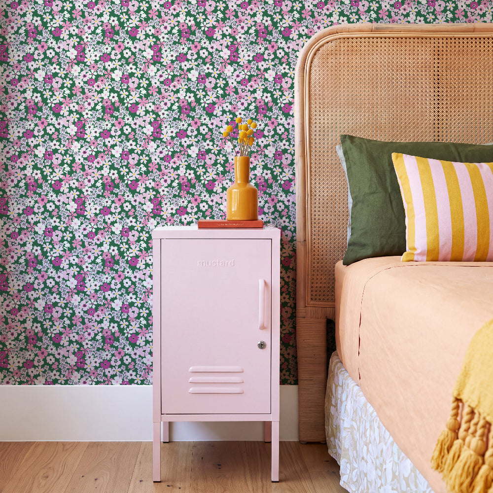 A pink metal nightstand in a bedroom setting, styled with a small yellow vase and flowers on top. The background is a vibrant green floral wallpaper, next to a bed with a woven cane headboard, green and yellow striped pillows, and peach-colored bedding.