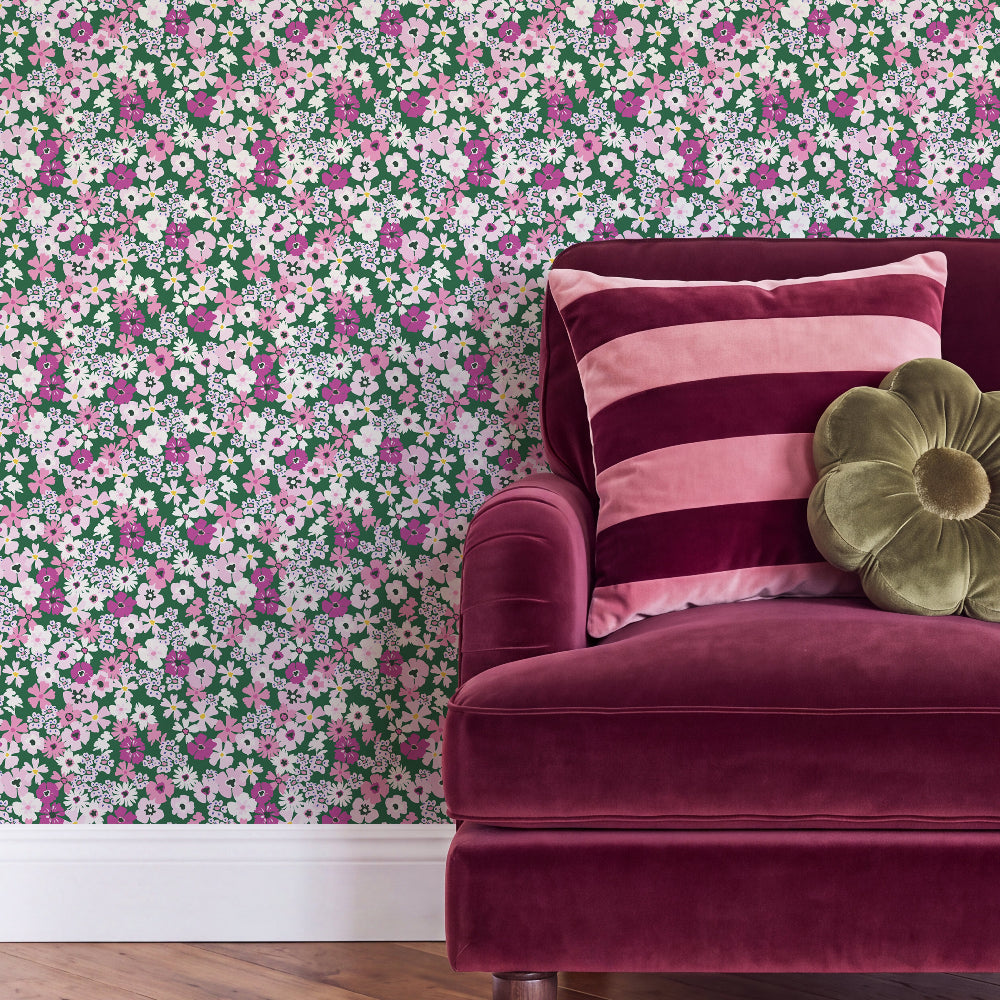 A close-up of a room corner featuring floral wallpaper with pink, white, and green flowers, styled with a deep burgundy velvet armchair, a pink striped throw pillow, and a green flower-shaped cushion.