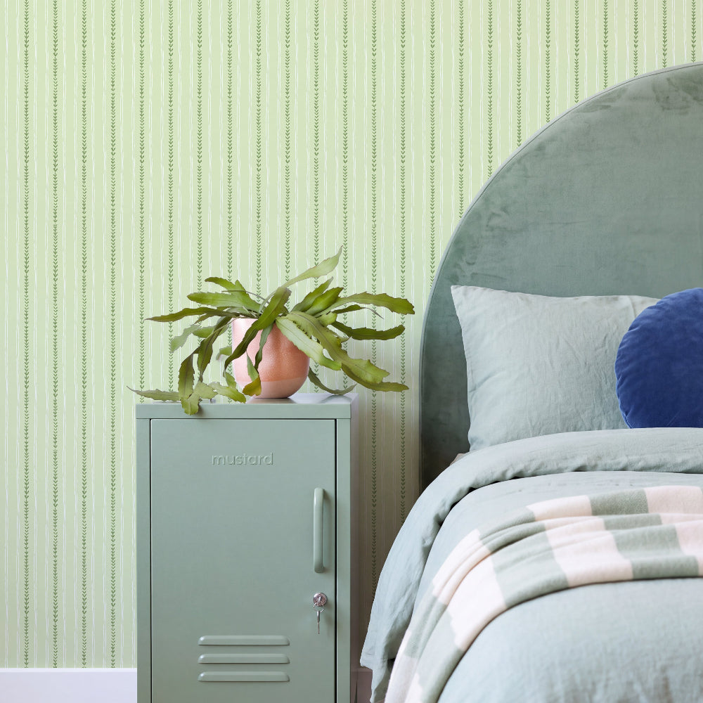 A styled bedroom scene featuring a sage green metal nightstand with a small potted plant on top, set against a light green vertically striped wallpaper. The nightstand is positioned next to a bed with a large round headboard, sage green bedding, and a blue accent pillow.