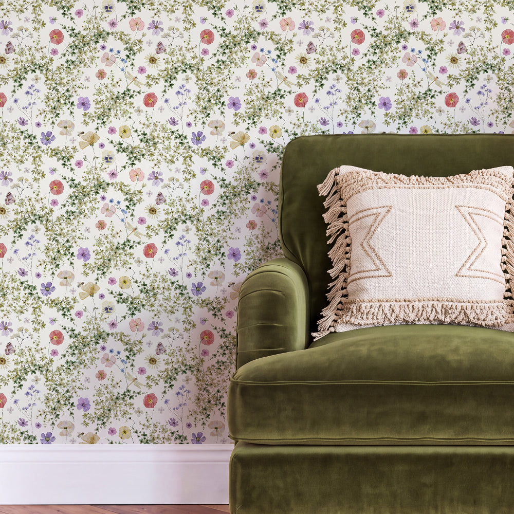 A close-up of a green velvet armchair with a cream fringed throw pillow, set against a wall covered in delicate multicolor floral wallpaper.