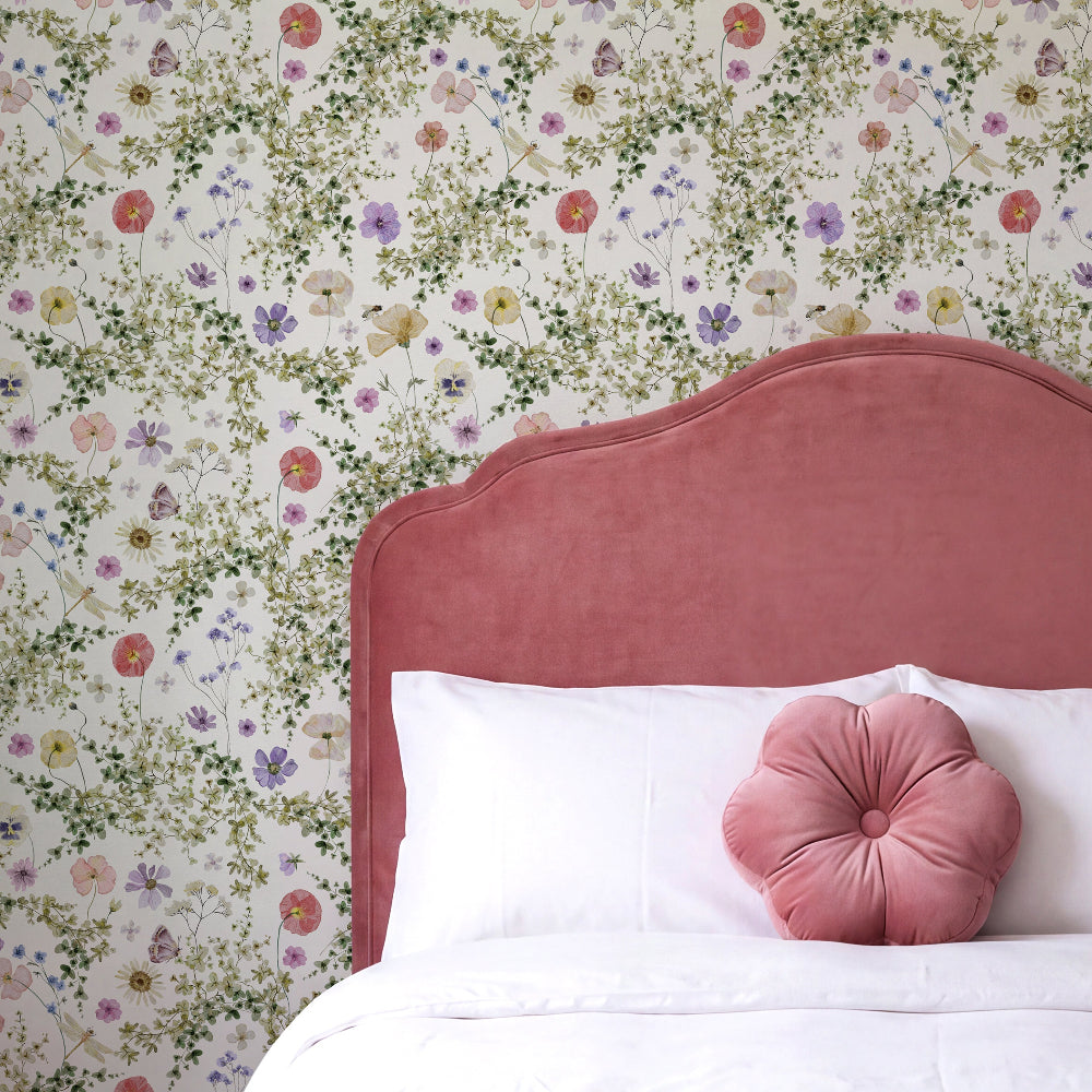 A close-up of a bed styled with a dusty rose velvet headboard, white bedding, and a matching pink flower-shaped pillow, set against a wall covered in a dense multicolor floral wallpaper.