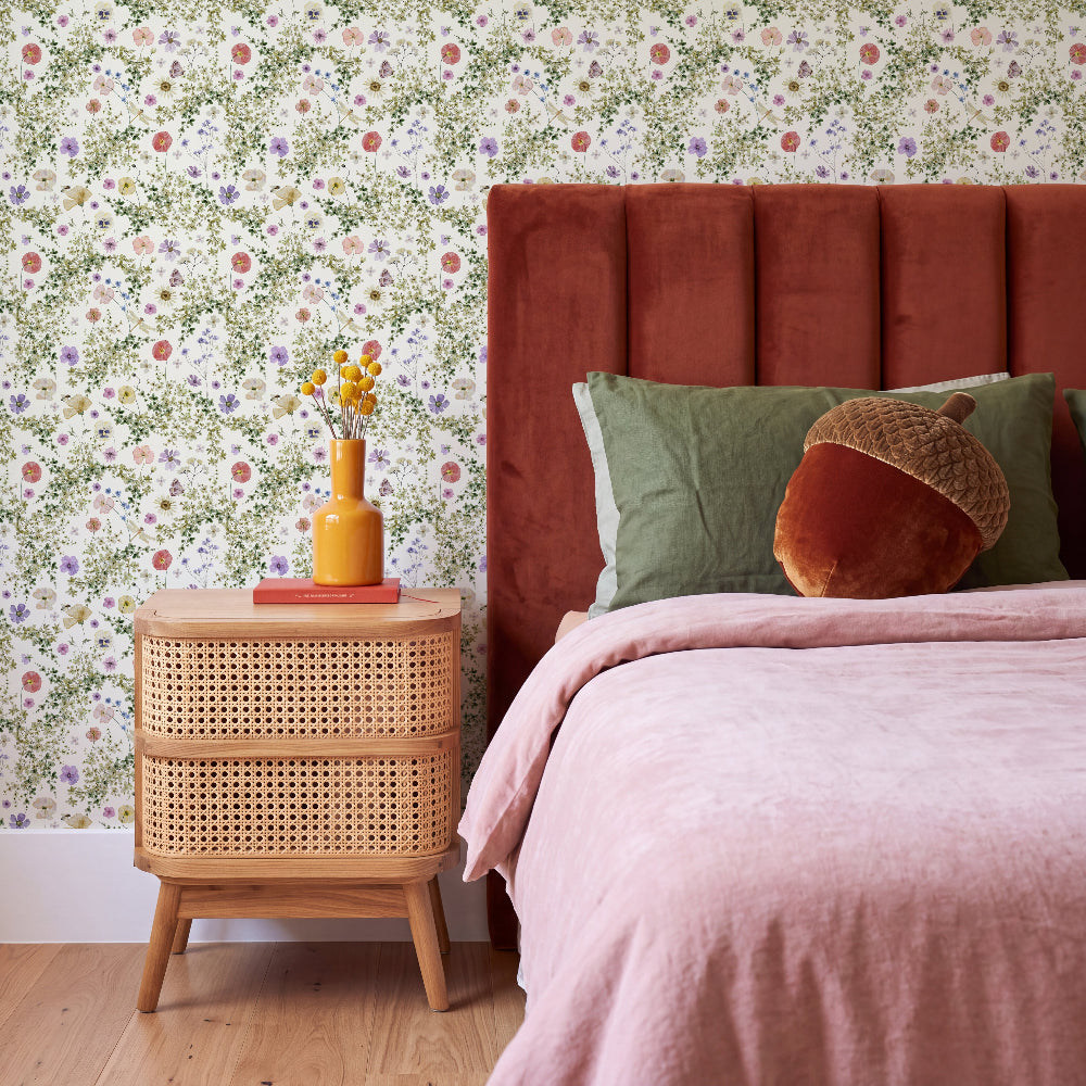 A styled bedroom corner featuring floral wallpaper, a rust-colored velvet headboard, pink bedding, a light wood cane nightstand, and a yellow vase with flowers.