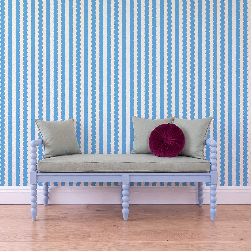 A light blue wooden bench with a pale sage green cushion and two pillows, including a round deep magenta accent pillow, set against a blue and white vertically striped wallpaper and light wood flooring.