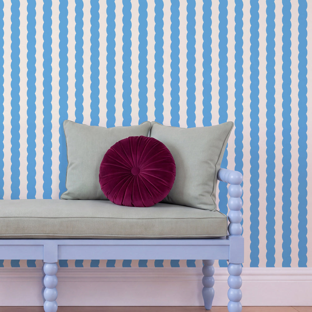 A styled interior scene showing a pale blue bench with a light beige cushion and two pillows, including a round deep magenta velvet accent pillow, set against a wall with blue and white wavy vertical stripes.