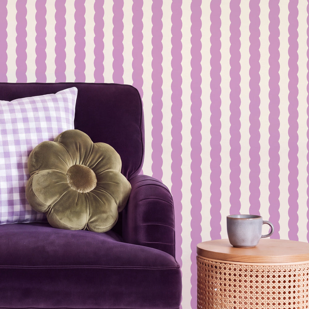 A styled interior scene showing a purple armchair with a green flower-shaped pillow and a white gingham pillow, set against pink and cream wavy vertical striped wallpaper. A small woven side table with a gray ceramic mug sits to the right.