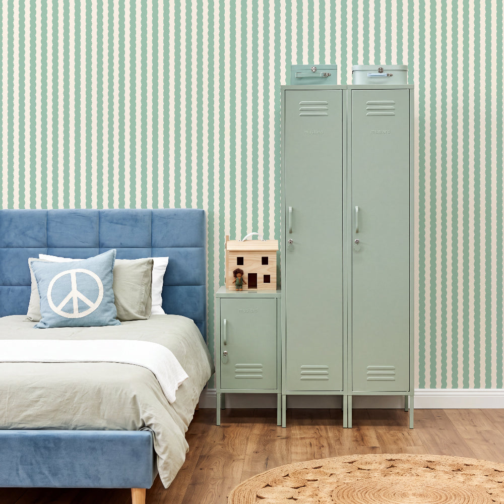 A styled bedroom scene featuring a tall mint green metal double-door locker and a matching side table next to a blue upholstered bed. The room has green and white vertically striped wallpaper, light wood floors, and a round woven rug.