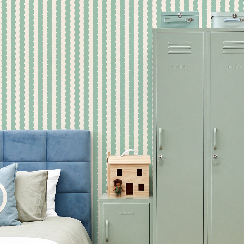 A styled bedroom corner featuring mint green and white wavy vertical striped wallpaper, a blue tufted headboard, a light green locker, and a small wooden house toy on a matching nightstand.