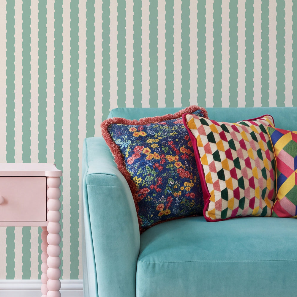 A styled interior scene showing a turquoise sofa with three decorative throw pillows, set against a mint green and white vertically striped wallpaper. A light pink side table is partially visible on the left.