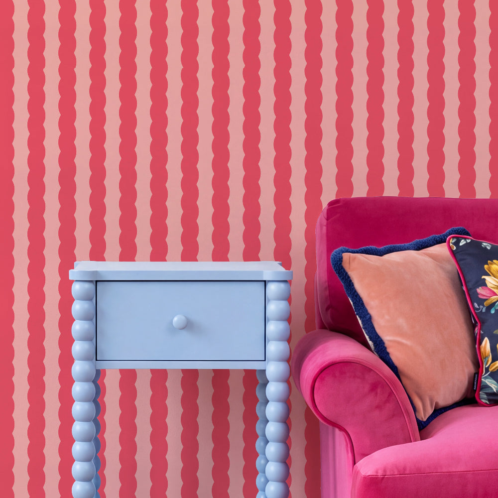 A styled interior corner showing a light blue bead-legged nightstand beside a bright pink armchair with decorative pillows, set against pink and coral wavy striped wallpaper.
