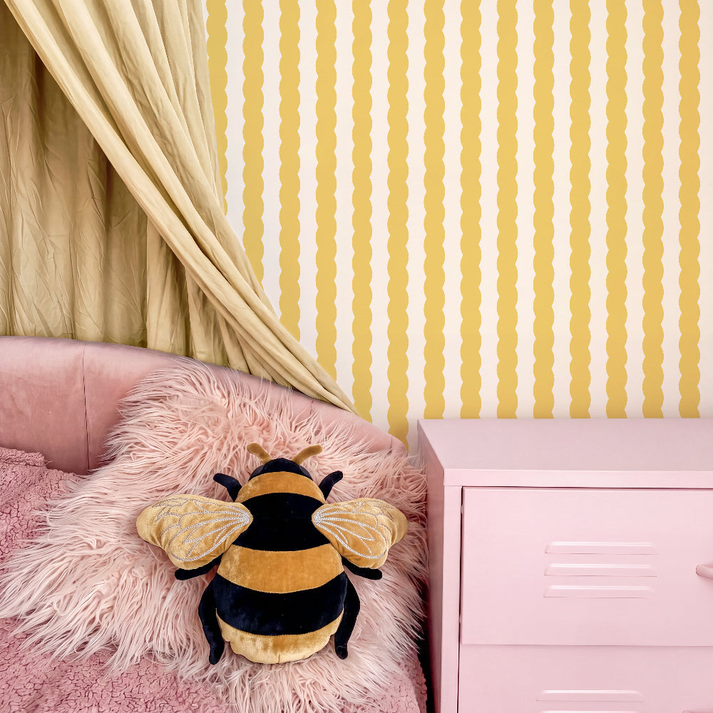 A plush bumblebee toy resting on a pink faux-fur pillow on a pink bed, with yellow and white wavy-striped wallpaper, a beige curtain, and a pink nightstand in the background.