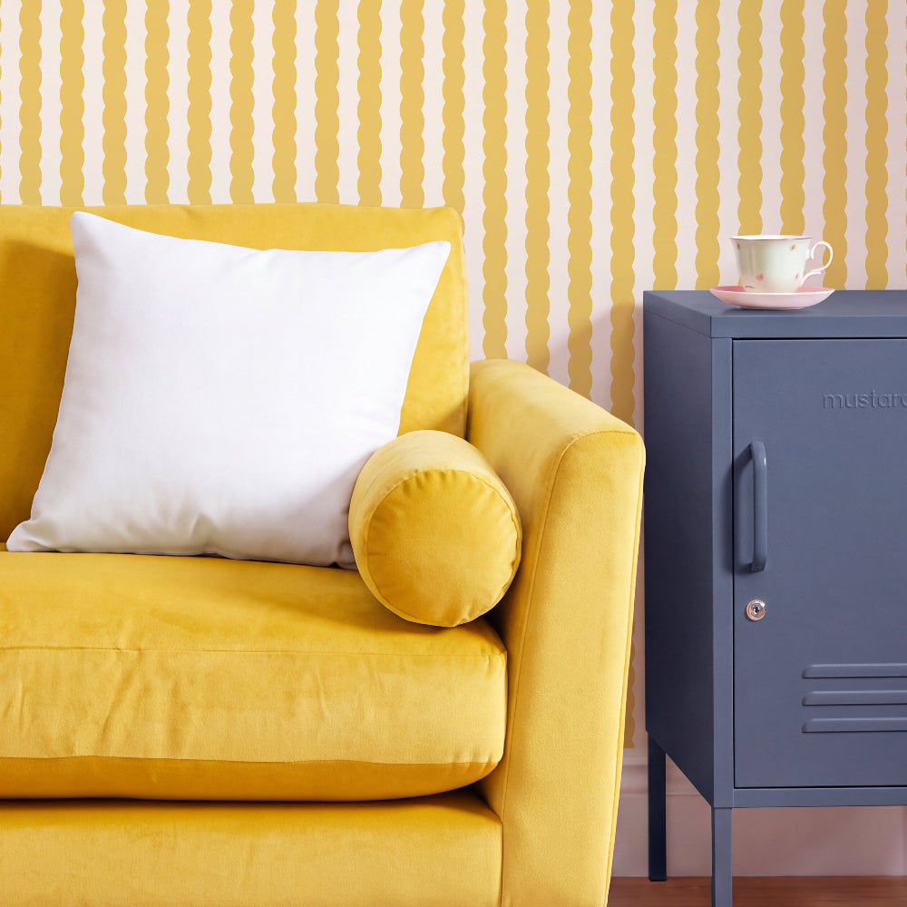 A yellow velvet sofa with a white throw pillow and a round yellow bolster cushion, set against a yellow and white wavy striped wallpaper. A dusty blue metal side cabinet with a teacup on top stands to the right.