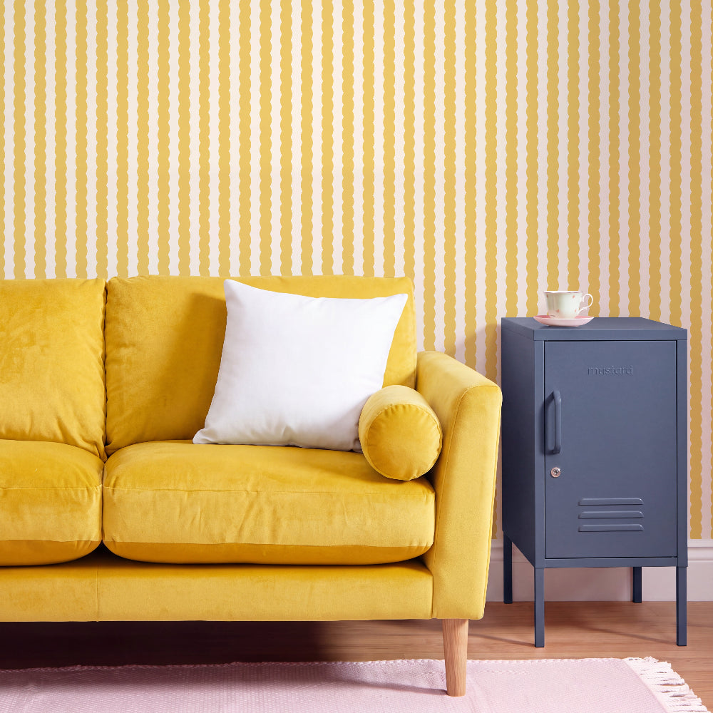 A yellow velvet sofa with a white throw pillow and a round yellow bolster pillow, set against a yellow and white wavy vertical striped wallpaper. A dark blue metal side cabinet stands to the right on a light wood floor with a pink rug.
