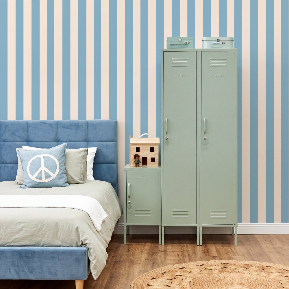 A styled bedroom scene featuring a tall mint green metal locker and a matching small side table next to a blue upholstered bed. The room has blue and cream vertical striped wallpaper, light wood flooring, and a round woven rug.
