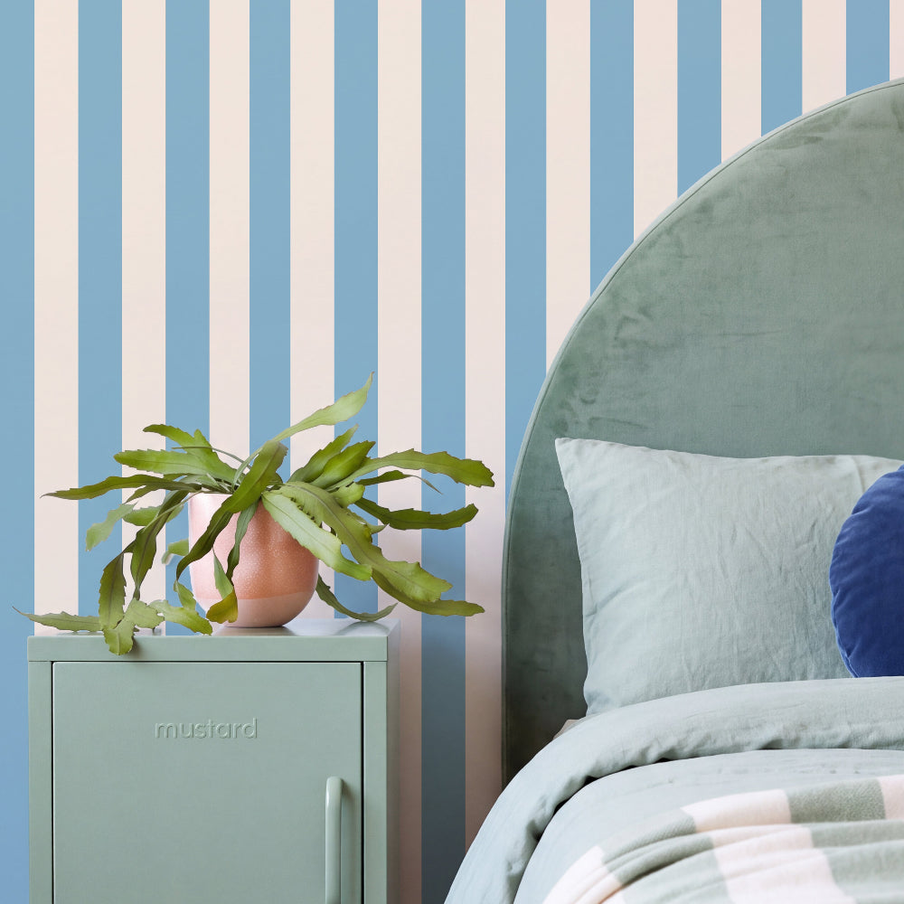 A styled bedroom corner featuring a mint green side table with a potted green plant, a light green velvet headboard, and a blue and white striped wallpaper background.