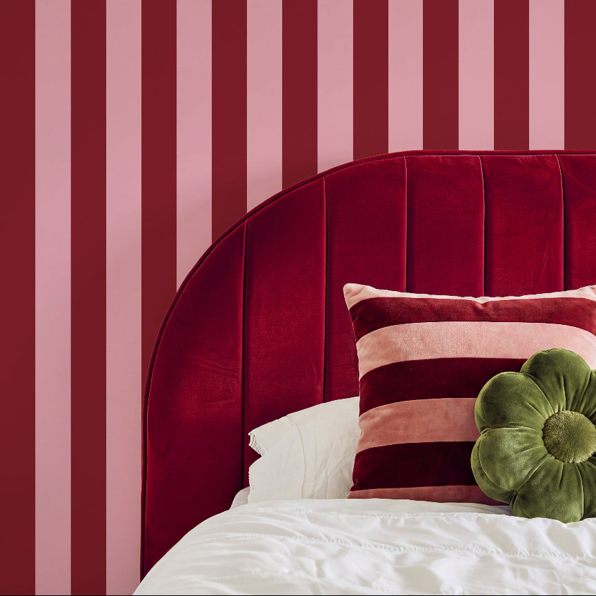 A styled bedroom scene featuring a bed with a deep red velvet headboard, white bedding, a striped pink and burgundy pillow, and an olive green flower-shaped cushion, set against pink and red vertically striped wallpaper.
