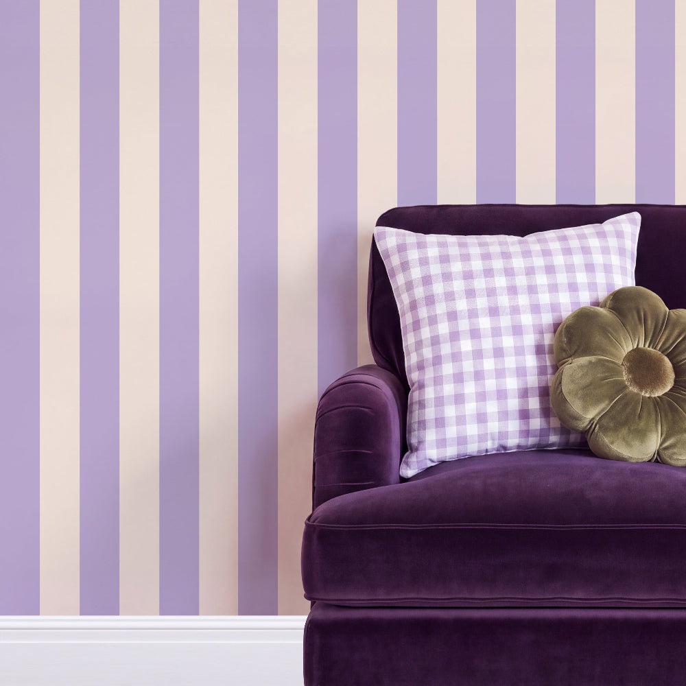 Purple and cream vertical striped wallpaper shown on a wall behind a purple velvet armchair with a lavender gingham pillow and a gold flower-shaped cushion.