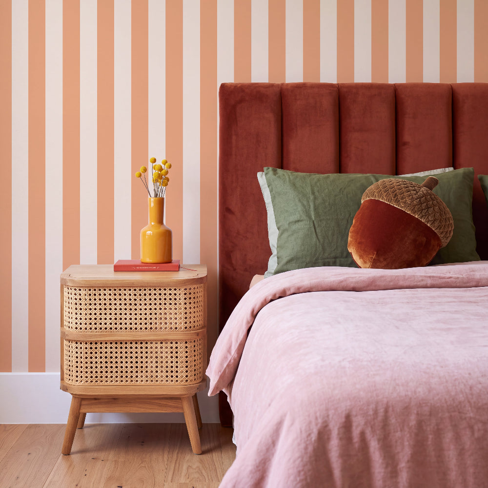 A styled bedroom corner featuring a bed with pink bedding, a tall rust-colored velvet headboard, a rattan nightstand, and striped peach and cream wallpaper.