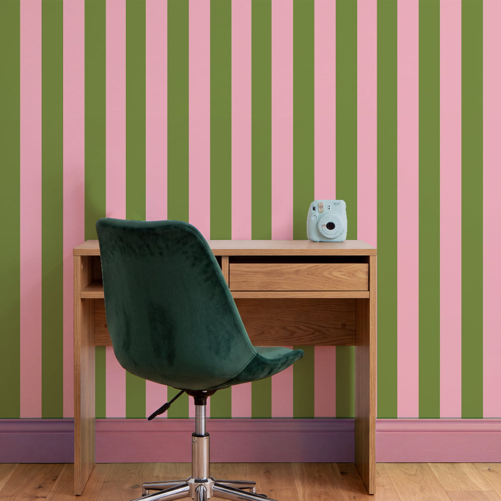 A styled interior scene with vertical pink and olive green striped wallpaper, a light wood desk, a dark green velvet swivel chair, and a small light blue instant camera on the desk.
