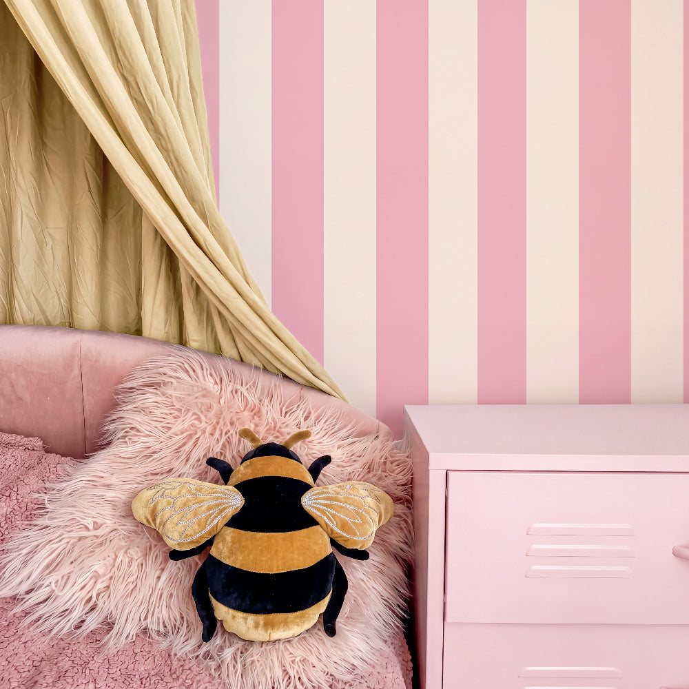 A plush bumblebee toy resting on a pink faux-fur pillow on a pink bed, with pink and cream striped wallpaper, a gold curtain, and a pink nightstand in the background.