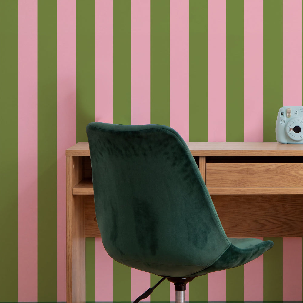 A dark green velvet swivel chair in front of a light wood desk, set against a wall with vertical pink and olive green stripes. A light blue instant camera sits on the desk.