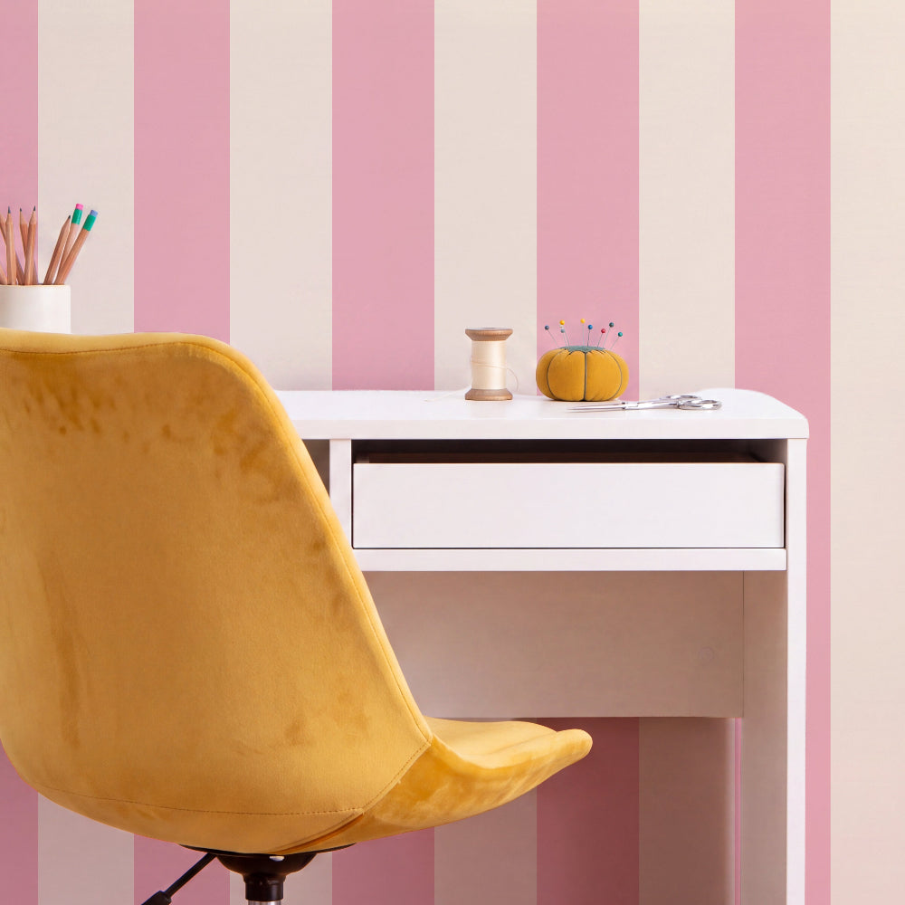 A yellow velvet office chair in the foreground, facing a white desk against a pink and cream vertically striped wall. A spool of thread, a small orange pincushion with pins, and a cup of colored pencils are on the desk.