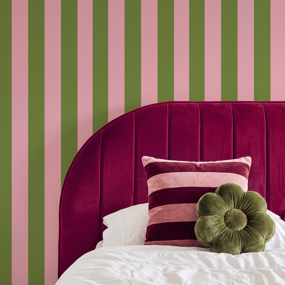 A styled bedroom corner featuring a bold vertical stripe wall in olive green and pink, with a large curved magenta velvet headboard, white bedding, a striped pink and burgundy pillow, and an olive green flower-shaped throw pillow.