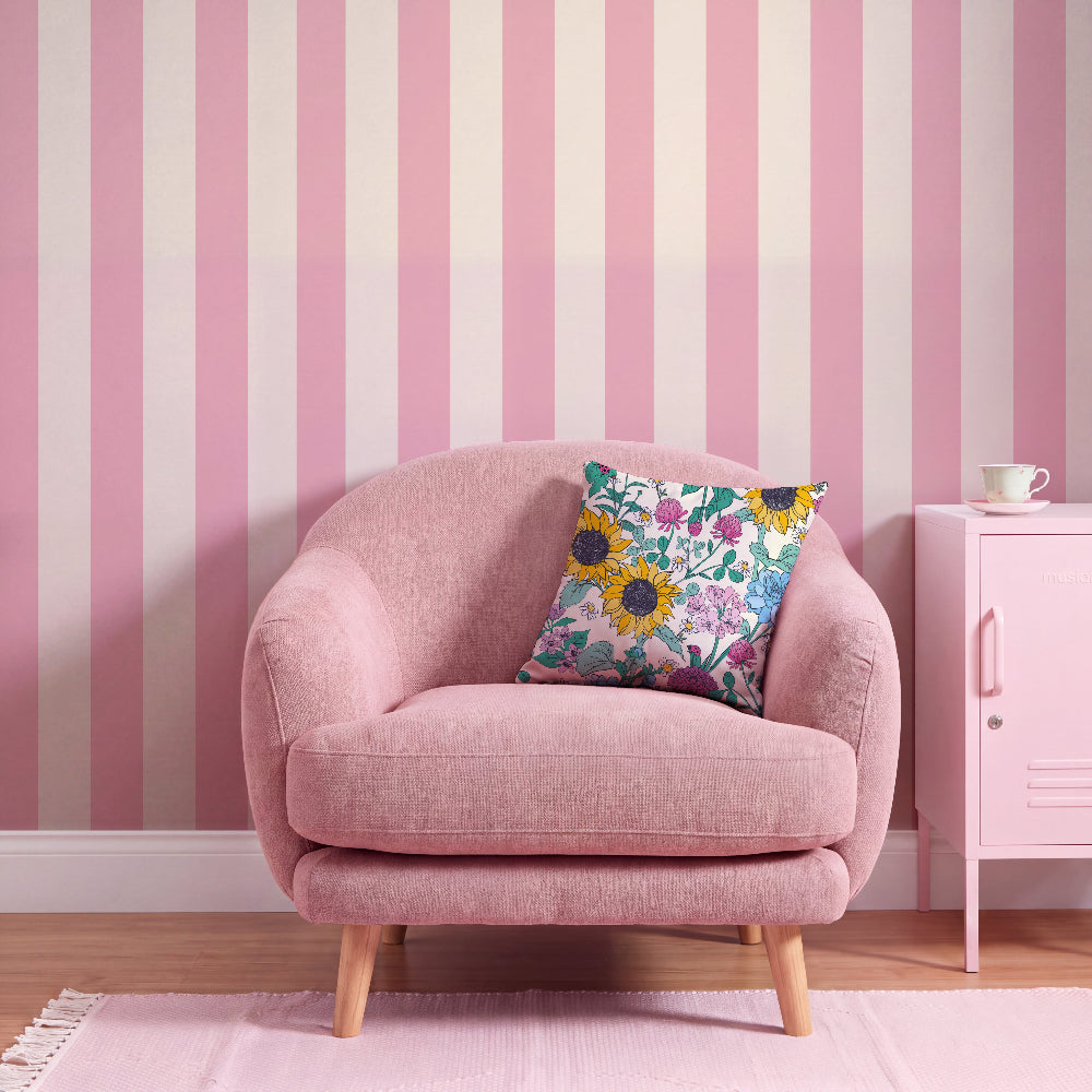 A pink upholstered armchair with light wood legs sits in a styled room against a pink and white striped wall, with a floral throw pillow on the chair and a small pink side cabinet to the right.