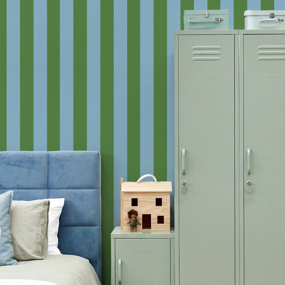 A styled bedroom corner featuring vertical green and light blue striped wallpaper, a blue upholstered bed headboard, a mint green metal locker, and a small wooden dollhouse on a matching nightstand.