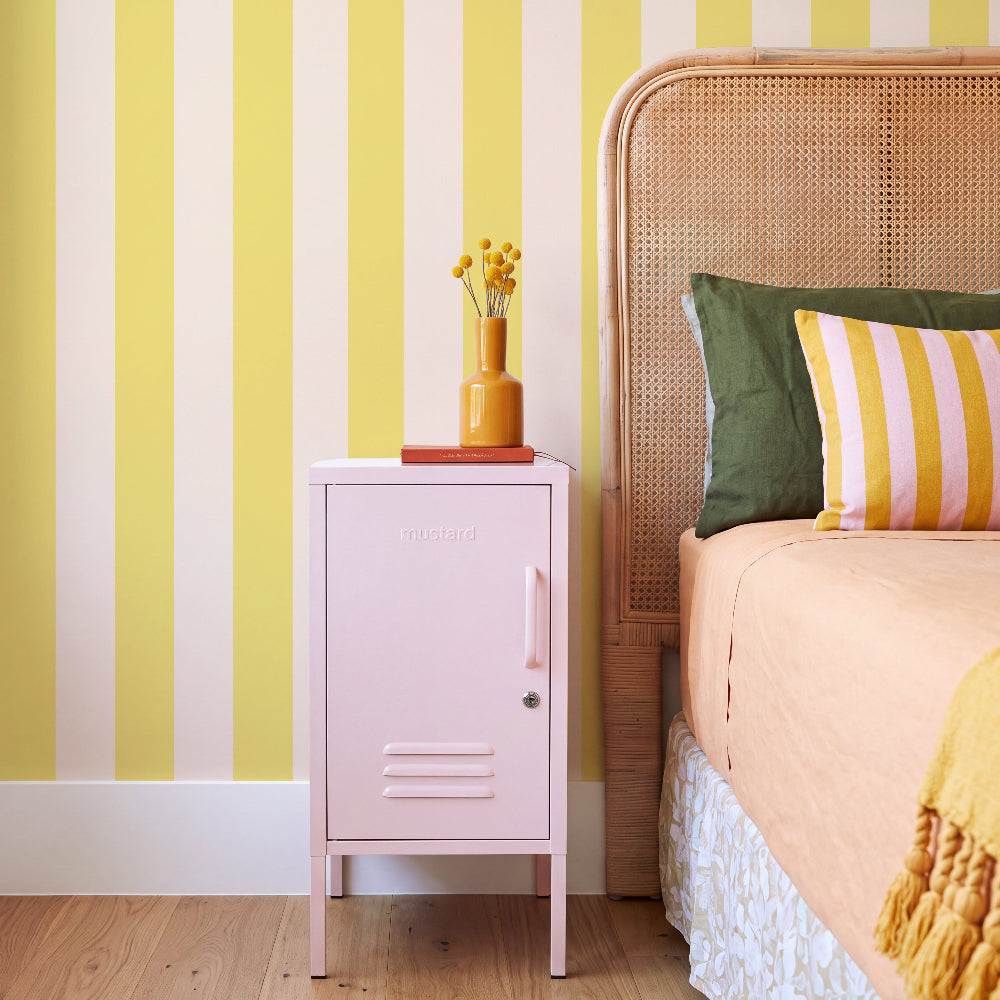A pink metal nightstand with a lock and handle stands beside a bed with a woven cane headboard in a bedroom. The wall has bold yellow and white vertical stripes, and the nightstand holds a small yellow vase with flowers and a red book.