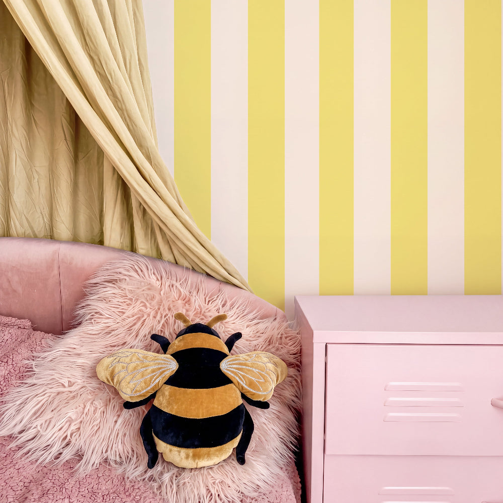 A plush bumblebee toy pillow with yellow and black stripes and translucent wings, resting on a pink faux-fur pillow on a pink bed in a styled bedroom setting with yellow and white striped wallpaper.