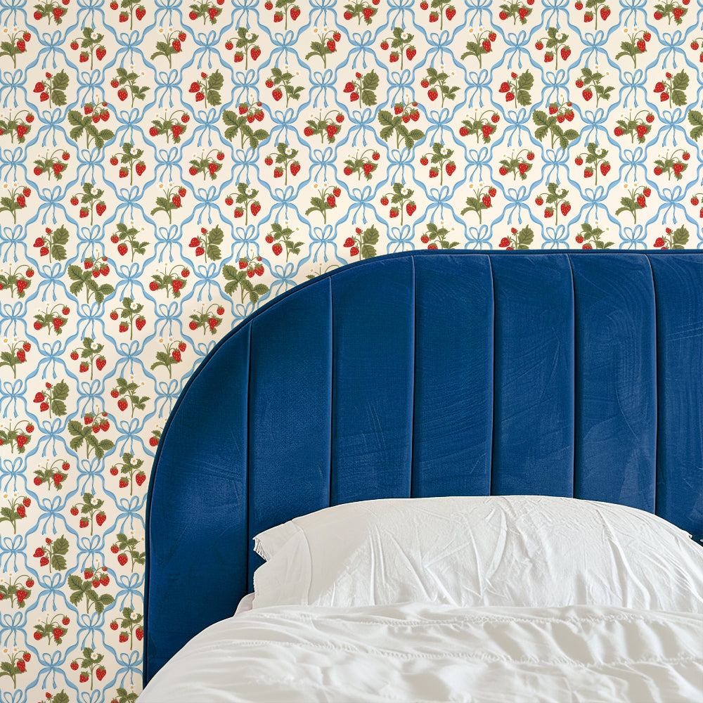 A close-up of a bed with a white pillow and rumpled white bedding in front of a blue upholstered headboard, set against wallpaper with a repeating strawberry and blue bow pattern.