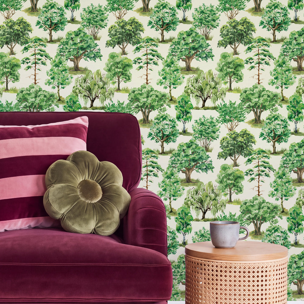 A styled interior scene showing a burgundy velvet armchair with pink striped and flower-shaped pillows, a small round wooden side table with a gray mug, and a wall covered in green tree-patterned wallpaper.