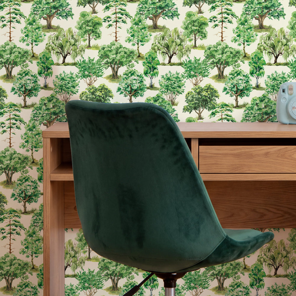A dark green velvet office chair positioned in front of a wooden desk, set against a wall covered in a repeating green forest tree wallpaper pattern.